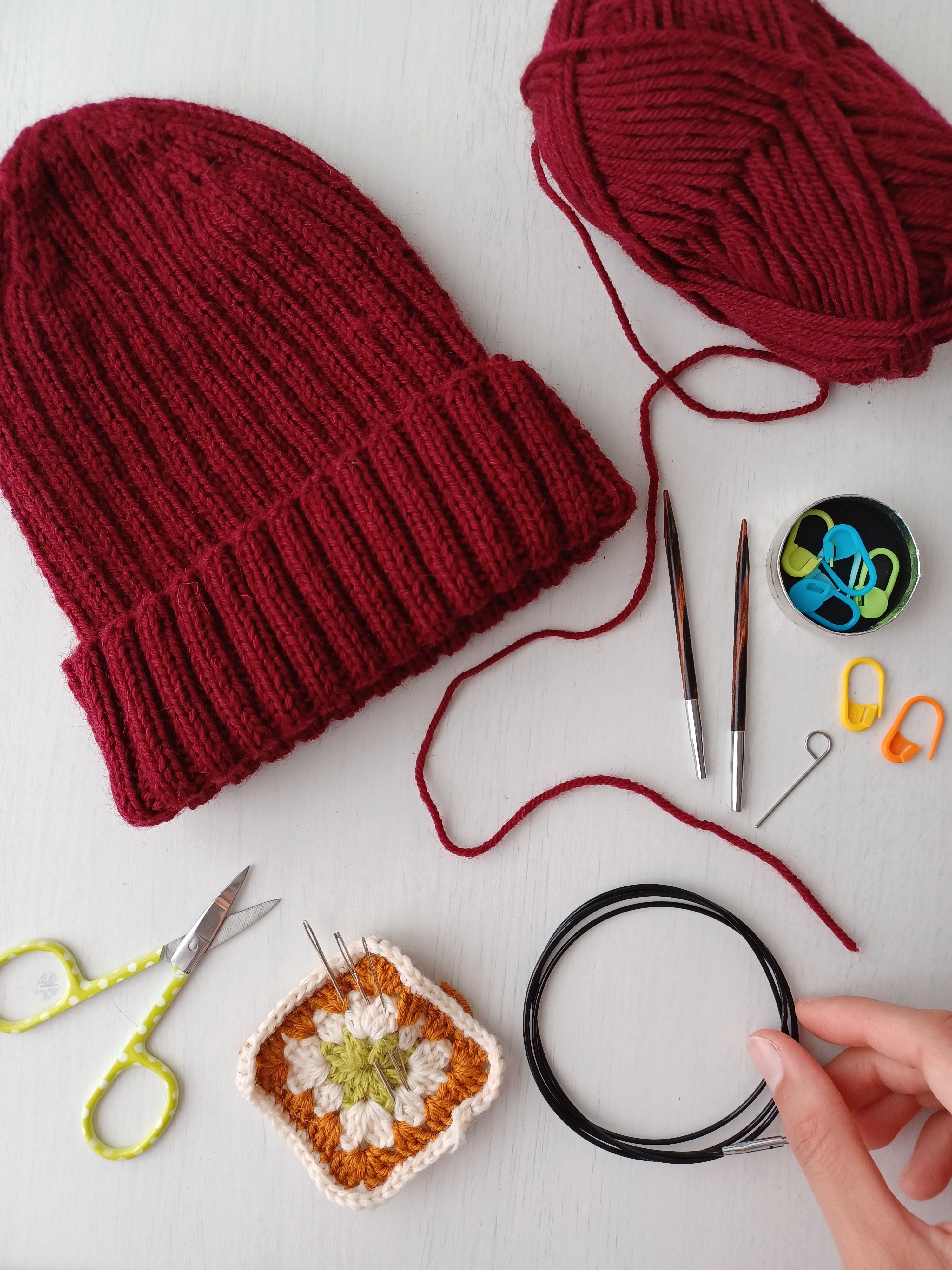 Un berretto a coste realizzato a maglia con un filato rosso ha accanto il gomitolo di filato con cui è stato realizzato e gli altri materiali necessari: segnamaglie, punte dei ferri circolari, cavo per i ferri circolari, ago da maglia e forbici