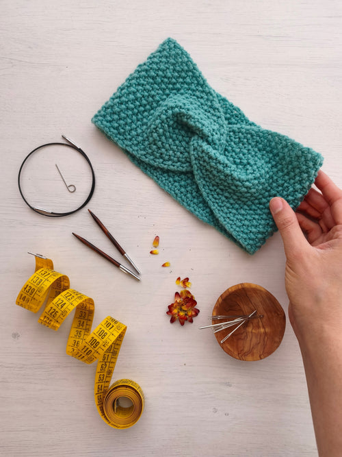 Una fascia azzurra realizzata a maglia con la tecnica del punto riso è appoggiata su un tavolo con accanto il cavo dei ferri circolari, le punte dei ferri, degli aghi da maglia e un metro da sarta