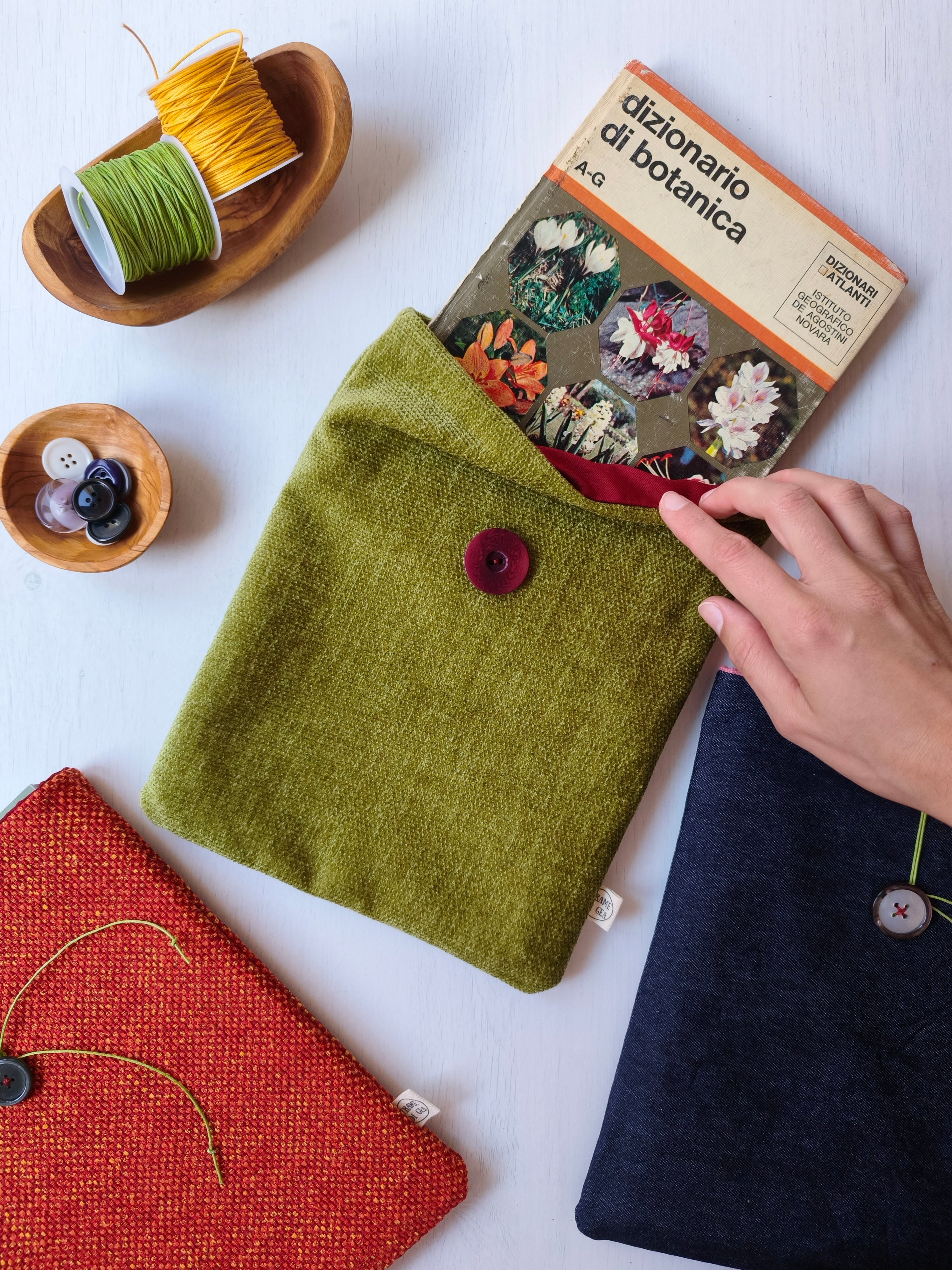 Tre custodie per libri di colori verde, rosso e blu con due rocchetti di fili e una ciotolina con dei bottoni
