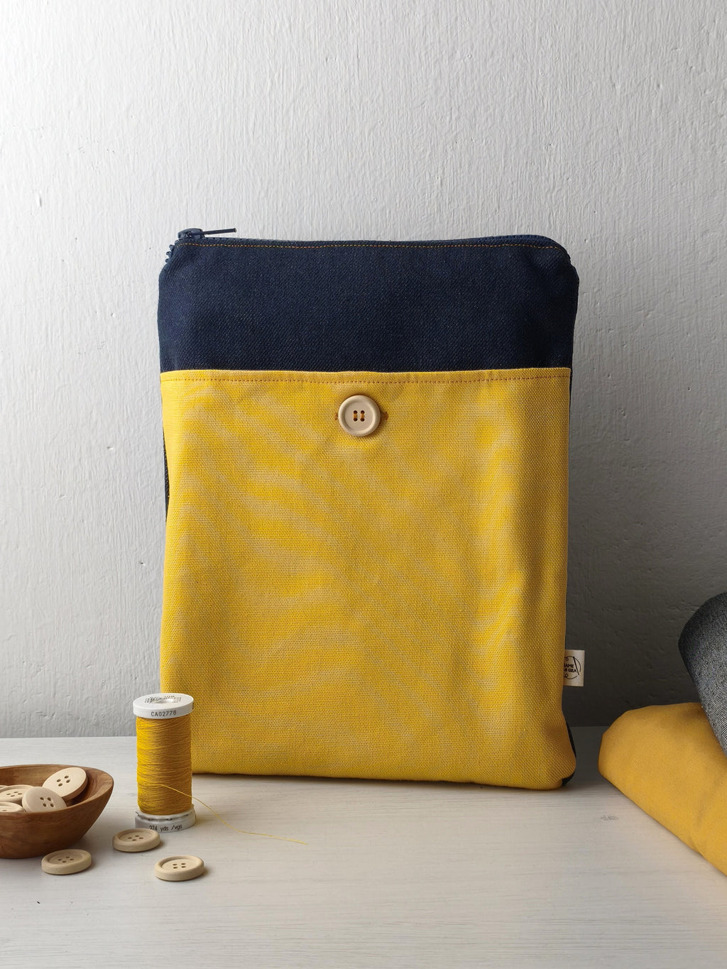Custodia per libri appoggiata sul tavolo con lampo, denim scuro e tasca gialla con bottone in legno