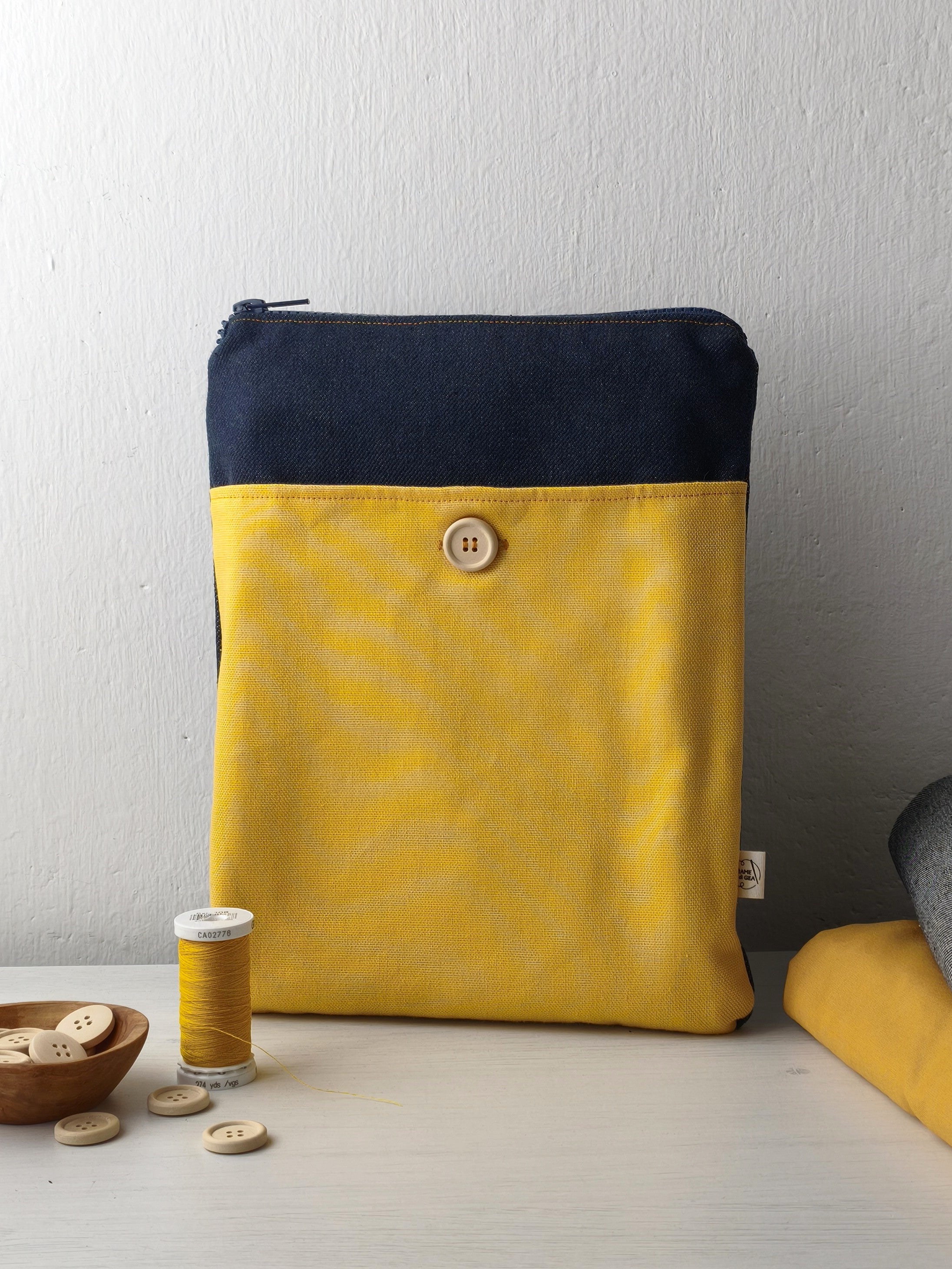 Custodia per libri appoggiata sul tavolo con lampo, denim scuro e tasca gialla con bottone in legno