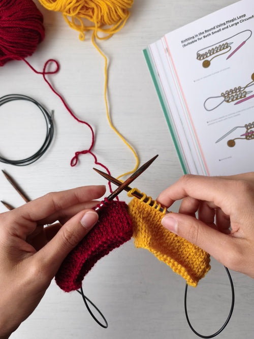 due mani tengono dei ferri da maglia circolari su cui sono state lavorate due circonferenze una di colore giallo e una di colore rosso, sullo sfondo un libro mostra come impostare la lavorazione del magic loop