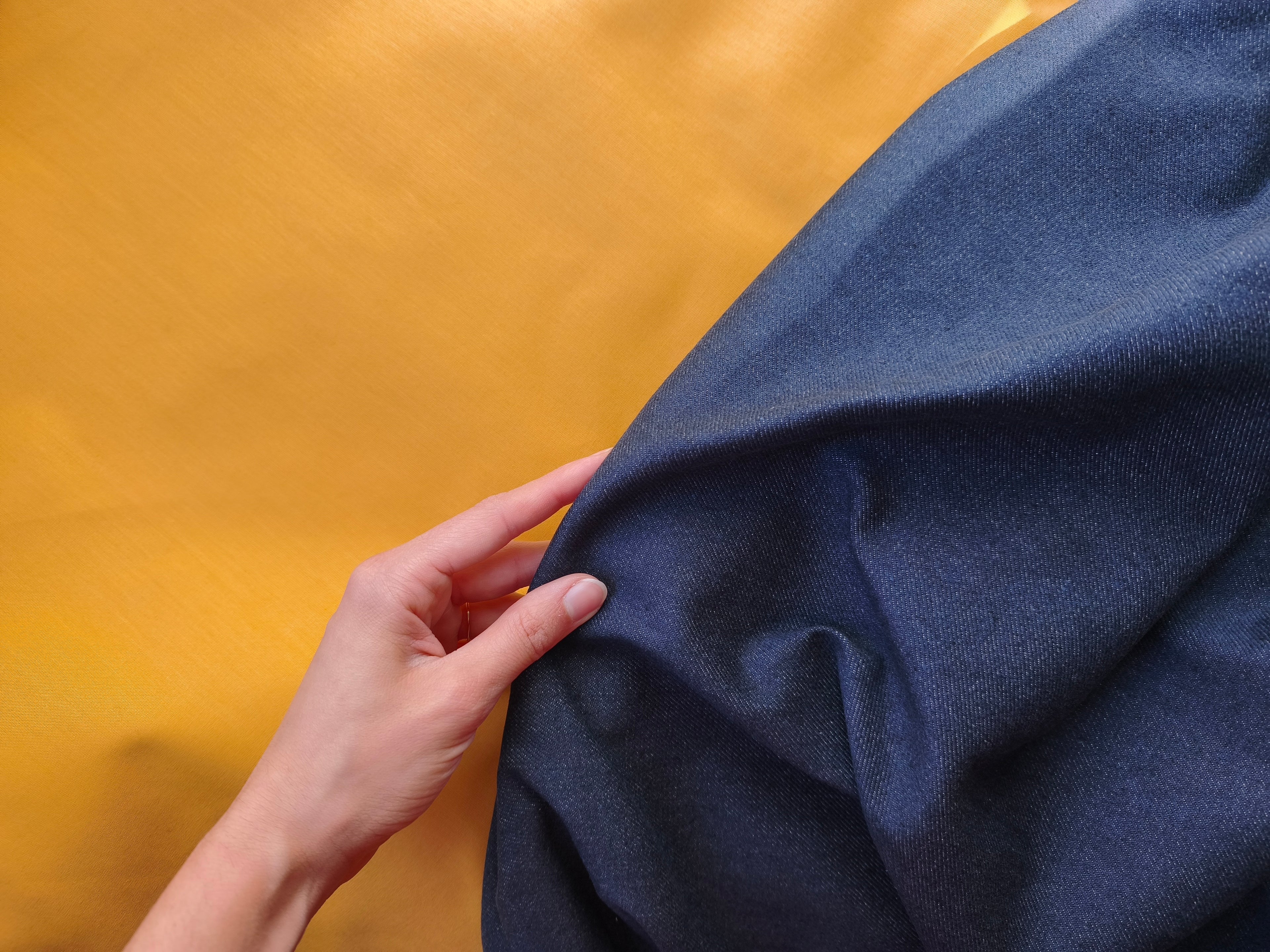 stoffa di jeans scuro che viene tirata con la mano sinistra sopra a una stoffa di cotone gialla, sono le stoffe della collezione primavera 2026
