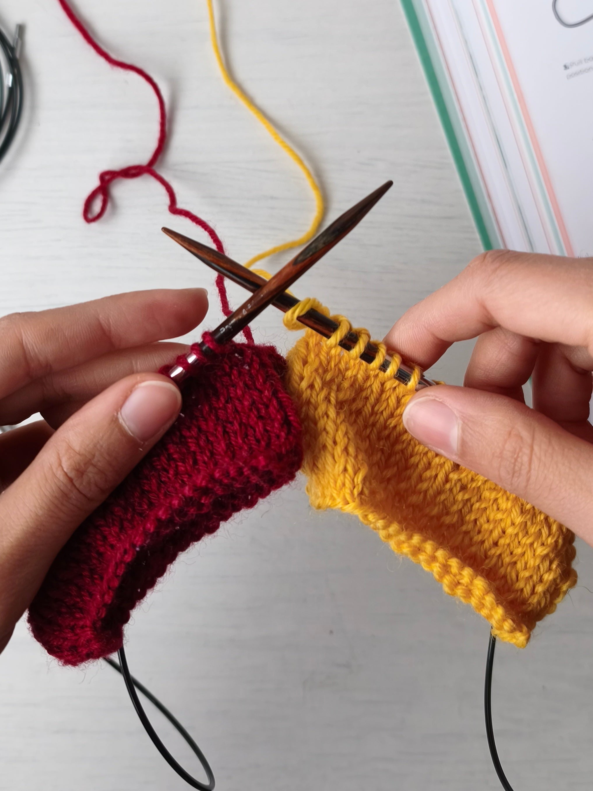 mani che tengono dei ferri da maglia  circolari su cui si stanno lavorando due circonferenze una di colore giallo e una di colore rosso, mostrano la tecnica del double magic loop